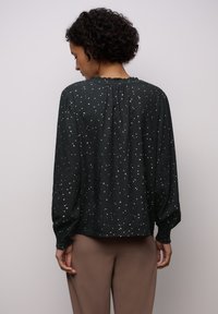 Tummanvihreä paita, jossa on hopeapilkullinen kuvio, laskostettu kaula-aukko ja pitkät hihat, joissa on joustavat hihansuut. Kevyt kangas, jossa on pehmeä tekstuuri.