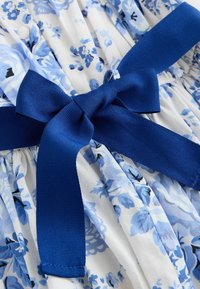 Tamsiai mėlyna audinio juosta surišta ant balto suknelės su mėlyna gėlėta spauda ir susmulkinto liemens detale.