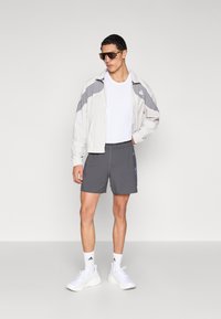 Light grey windbreaker with dark grey accents, white t-shirt, dark shorts with light blue stripes, and white athletic shoes. Brown sunglasses.