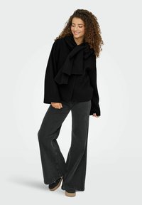 Sort langærmet sweater med en stor sløjfe ved halsen, parret med mørkegrå vide bukser og sorte sko med en struktureret sål.