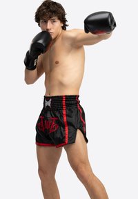 Sorte bokseshorts med røde accenter og et grafisk design foran, parret med sorte boksehandsker, model i en kampstilling.