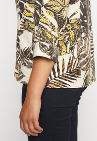 Kortærmet bluse i let stof med et livligt tropisk bladprint i gult, brunt og sort, med opsmøgede manchetter og en afslappet pasform.