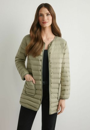 Kobieta z długimi brązowymi włosami nosi jasną zieloną pikowaną kurtkę z zatrzaskami na czarnej bluzce i czarnych spodniach, stoi na jednolitym tle.