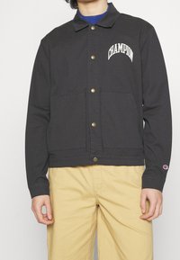 Czarna kurtka coacheska z logo na piersi i dwiema przednimi kieszeniami. Posiada złote napy, kołnierzyk oraz elastyczne mankiety.