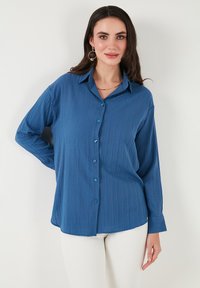 Camisa azul de manga larga con un tejido texturizado, con un cierre de botones en la parte delantera y botones a tono. Diseño con cuello y un ajuste relajado.