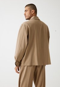 Giacca beige su misura con colletto a scollo, maniche lunghe e dettagli con bottoni, abbinata a pantaloni coordinati, presentata su uno sfondo neutro.