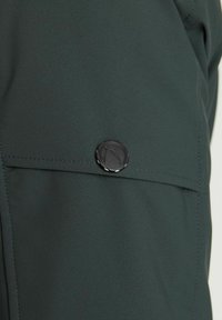 Tela verde oscuro mostrando un primer plano de un bolsillo con un botón redondo negro que presenta un logo, textura suave y detalle de costura prolijo.