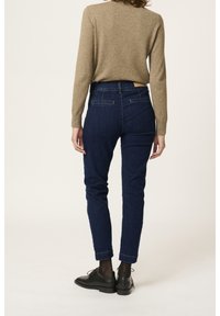 Jean bleu foncé à coupe fuselée et taille haute, associé à un pull marron clair ; dispose de poches arrière classiques et de détails minimaux.