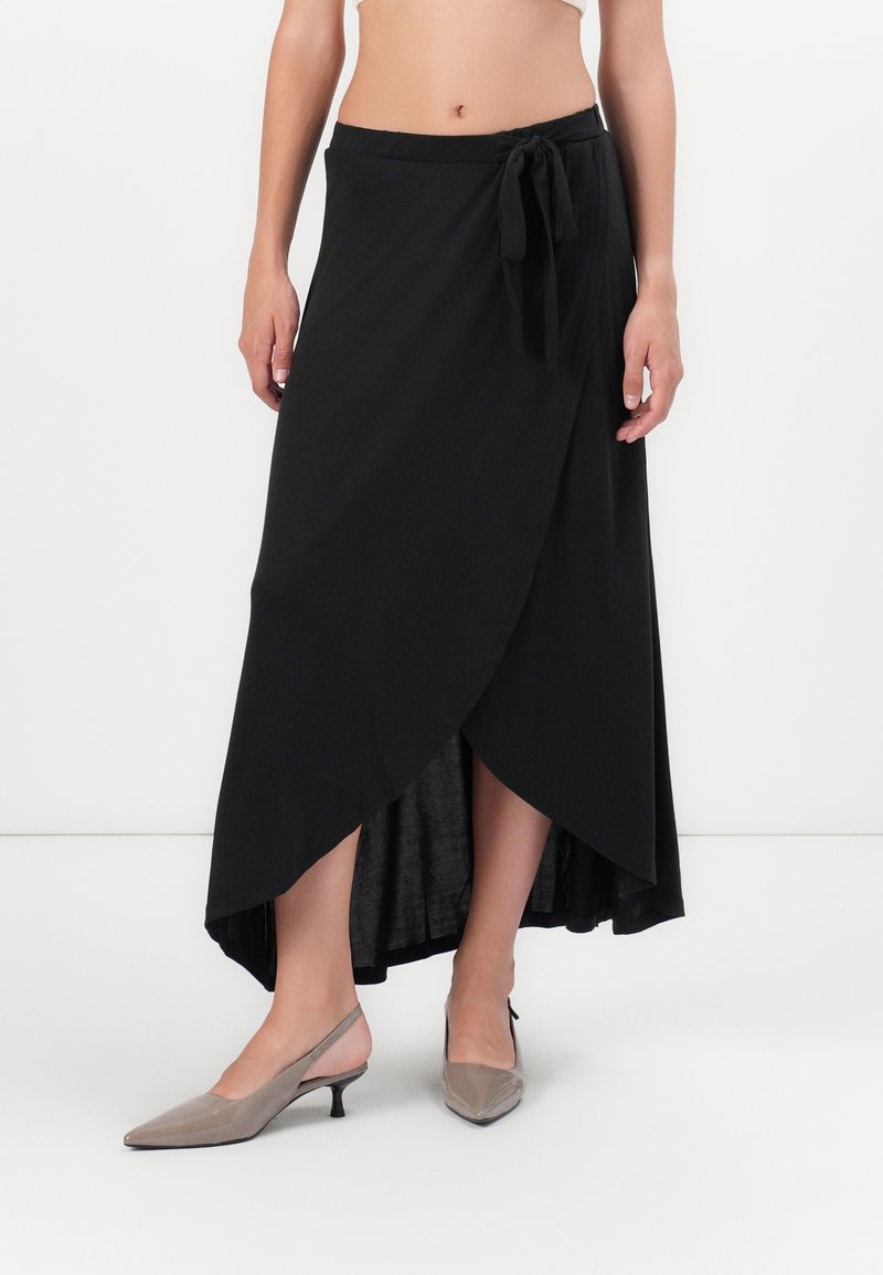 Jupe noire enroulée avec un lien à la taille, présentant un ourlet asymétrique. Tissu lisse avec un léger éclat, associé à des escarpins gris clair à lanières.