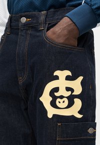 Σκούρο τζιν παντελόνι με κίτρινο γραφικό σχέδιο που απεικονίζει ένα στυλιζαρισμένο χαρακτήρα, πορτοκαλί ραφές και πλαϊνή τσέπη με λεπτομέρεια κουμπιού.