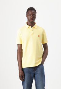 Žlté bavlnené polo tričko s krátkymi rukávmi, troj-buttonová légou a malé oranžové logo na ľavej hrudi. Nosené s modrými džínsami.