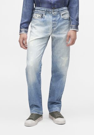Personne portant un jean bleu délavé avec une légère usure et des baskets basses grises, debout devant un fond clair uni.