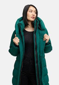 Giacca a vento verde lunga con collo alto, cerniere e bottoni. Texture liscia con un design trapuntato. Indossata sopra un top nero.