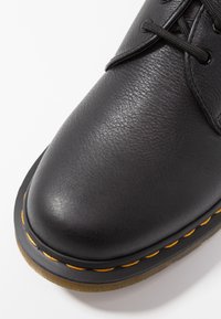 Scarpa in pelle nera con punta arrotondata, superficie texturizzata e cucitura arancione lungo il bordo della suola nera. I lacci sono neri con occhielli in metallo.