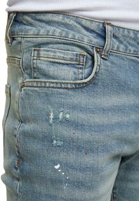 Jeans de mezclilla azul claro con parches desgastados y pequeñas manchas de pintura roja y blanca en la parte del muslo delantero.