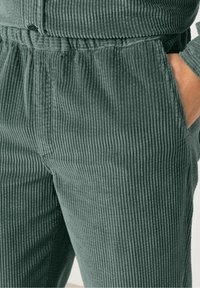 Pantalone iz kordura v umirjeni zeleni barvi, z vertikalno ribano teksturo, elastičnim pasom in stranskimi žepi.