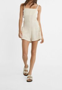 Shorts salopette in lino beige con bretelle regolabili, legature laterali e una vestibilità comoda, abbinati a sandali chiari con fibbie.