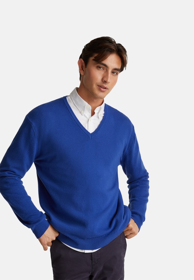 V-Ausschnitt, langärmliger Pullover in lebhaftem Königsblau, aus weichem Material mit gerippten Bündchen und Saum, über einem weißen Hemd mit Kragen getragen.