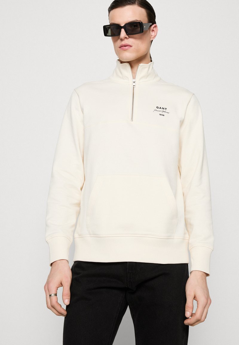 Felpa pullover color crema con zip a un quarto, tasca frontale e logo GANT. Realizzata in un morbido tessuto dalla superficie liscia.