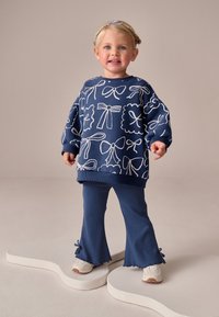 Sweat-shirt bleu marine avec imprimé nœud blanc, assorti à un pantalon évasé côtelé assorti. Les chaussures sont claires avec des accents latéraux. L'enfant se tient sur une surface lisse.