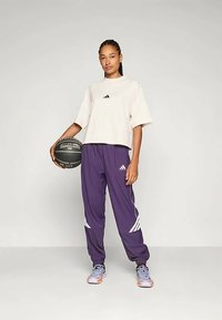 Cremfarbener kurzärmeliger Cropped-T-Shirt mit schwarzem Logo, kombiniert mit lila, schmal zulaufenden Hosen mit weißen Akzenten und grauen Turnschuhen, während er einen Basketball hält.