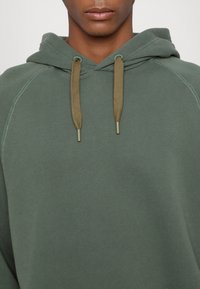 Felpa color verde oliva realizzata in tessuto morbido, con tasca a marsupio, cappuccio regolabile con coulisse e punte in metallo, e dettagli di cucitura a contrasto.