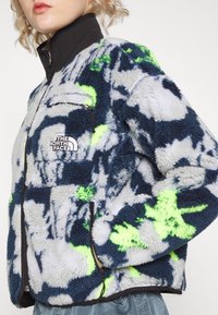 Szara kurtka polarowa zdobiona abstrakcyjnym wzorem w kolorze granatowym i neonowozielonym. Posiada zapinaną na zamek kieszeń na piersi oraz czarny kołnierz.