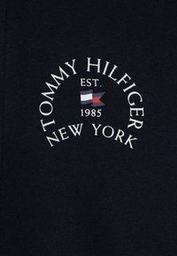 Jūros stiliaus audinys su baltu užrašu "TOMMY HILFIGER EST. 1985 NEW YORK". Papuoštas mažu raudonu ir mėlynu vėliavos emblematu. Lygus, minkštas paviršius.