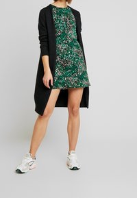 Robe verte à imprimé floral et à manches courtes, superposée sous un cardigan en tricot sombre. Le modèle porte des chaussures de sport blanches.
