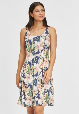 Vrouw draagt een mouwloze jurk tot op de knie met blauwe achtergrond en grote witte, perzikkleurige en groene bloemenprint, kijkt iets opzij.