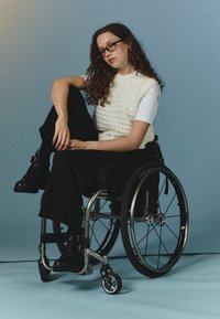 White knitted vest over a white shirt, black wide-leg pants, and black ankle boots. The individual is sitting in a wheelchair with a silver frame.