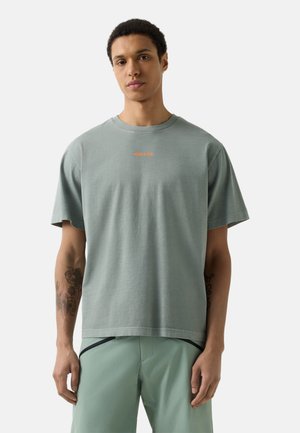 Junger Mann mit Tätowierungen, der ein locker sitzendes hellgraues T-Shirt mit orangefarbenem Text und passende hellgrüne Shorts vor einfachem Hintergrund trägt.