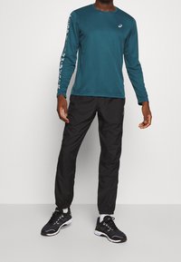 Langärmliges, türkisfarbenes Hemd mit weißem Logo auf dem Ärmel, kombiniert mit schwarzen, schmal geschnittenen Hosen und schwarzen Sportschuhen mit weißen Sohlen.