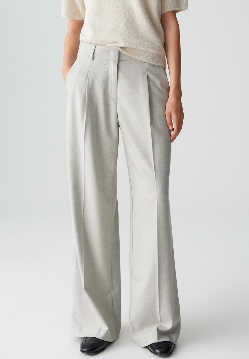 Pantalons larges taille haute gris clair avec des plis. Tissu lisse avec une coupe ajustée. Portés avec un pull ample de couleur crème.