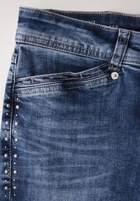 Jeans in denim blu con vestibilità slim, borchie ai lati e chiusura con bottone. Dettagli di scolorimento e cuciture evidenti lungo la vita e le tasche.
