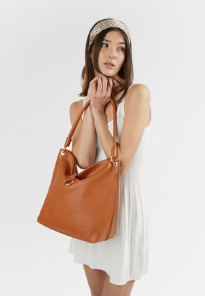 Giovane donna in abito bianco e fascia per capelli decorata che tiene una grande borsa a tracolla in pelle marrone con dettaglio ad anello dorato.