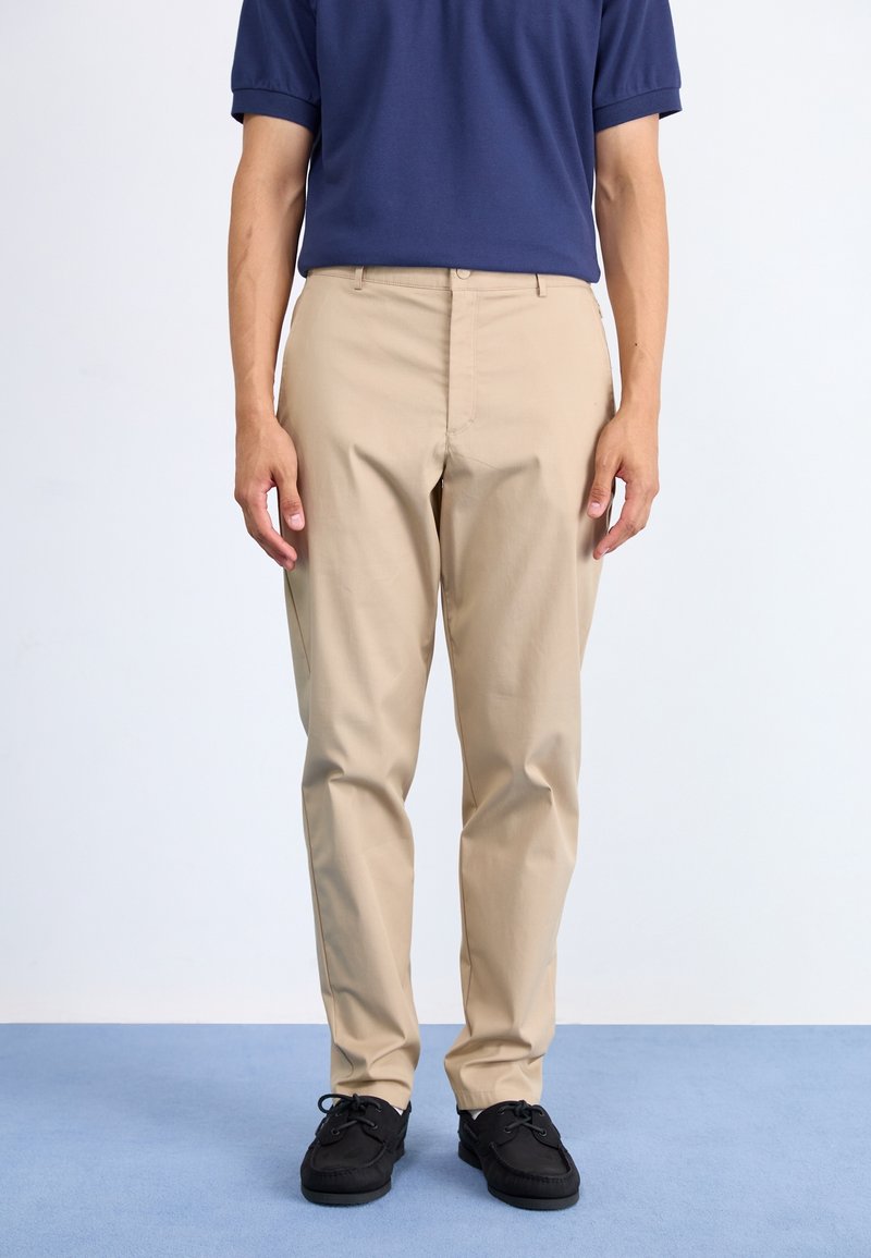Homme portant un pantalon chino beige, un polo bleu marine rentré à l'intérieur, et des chaussures noires à lacets, debout sur un tapis bleu avec un fond blanc.