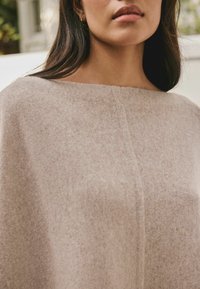 Beige uldponcho med en bred, firkantet halsudskæring, der har en struktureret overflade og et centralt sømdetalje. Blød, varm fremtoning.