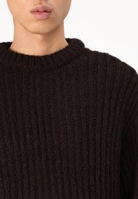 Mörkbrun stickad tröja med ribbad textur och rund halsringning. Tyget ser tjockt och varmt ut, med en något lös passform.
