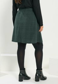 Grünes kariertes Kleid aus strukturiertem Stoff, mit einem ausgestellten Design und einem hohen Bund. Kombiniert mit schwarzen Strumpfhosen und Stiefeletten.