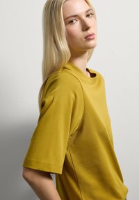 Top giallo senape a maniche corte con scollatura rotonda, realizzato in tessuto morbido. Presenta una vestibilità rilassata e una lavorazione delle cuciture minima.