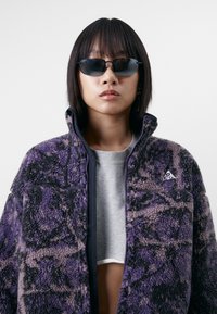 Giacca in fleece a motivo viola e nero con colletto alto, zip sul davanti e dettaglio del logo sul petto, indossata sopra un maglione grigio.