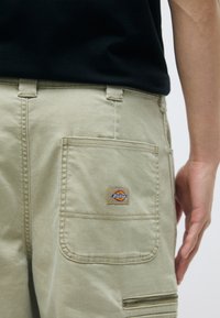 Béžové cargo nohavice vyrobené z odolného materiálu. Obsahujú veľkú zadnú kapsu s nášivkou a malú zipsovú kapsu na strane.