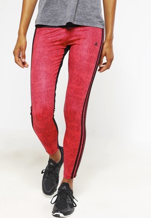 Leggings rouges à motifs avec des bandes latérales noires, fabriqués dans un matériau extensible, associés à des chaussures de sport noires et un haut gris.
