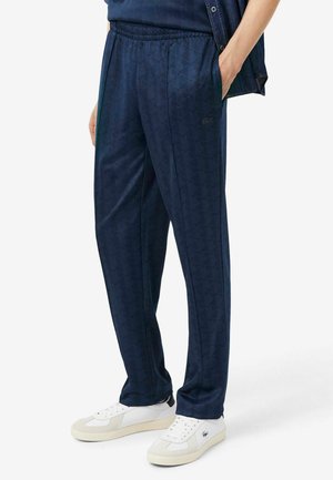 Person trägt marineblaue gemusterte Hose mit elastischem Bund und weiße Sneakers mit Logo vor einfarbigem Hintergrund.
