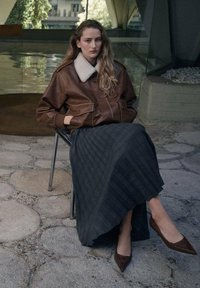 Chaqueta bomber de cuero marrón con cuello de piel, falda larga plisada gris y tacones marrones de punta, sentada en una silla de metal.