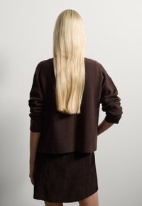 Suéter marrón de punto con mangas largas, combinado con una falda de pana marrón oscuro. Se ve el cabello rubio largo y liso.