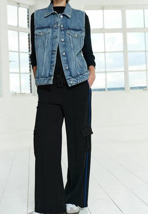 Džínová bezrukávová bunda na knoflíky, kombinovaná s černými širokými kalhotami typu cargo s modrými pruhy po stranách a několika kapsami.