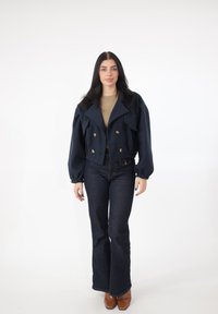 Granatowa dwurzędowa krótka marynarka z przednimi kieszeniami, bufkami na rękawach i złotymi guzikami, zestawiona z ciemnymi, rozszerzanymi jeansami i brązowymi butami.