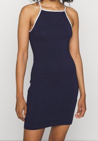 Marineblaues ärmelloses Kleid mit taillierter Silhouette, ausgestattet mit weißen Akzentträgern und einer weichen, glatten Textur.
